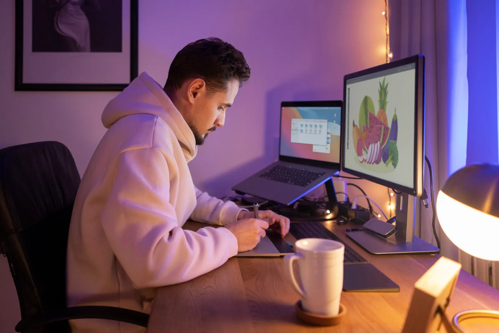 Un créateur web dessine sur une tablette graphique devant deux écrans, dans un bureau éclairé de lumières violettes